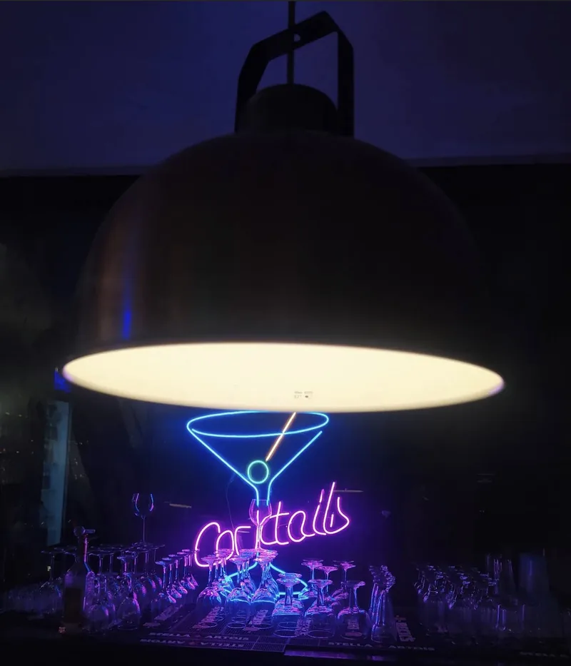 Une enseigne au néon « Cocktails » sous une grande lampe suspendue, dans le noir.